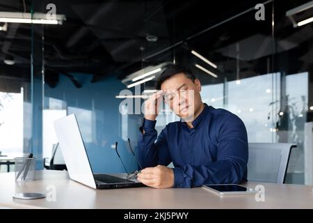 thinking sad asian man with closed eyes working in office indoors, businessman sitting at desk overworked and frustrated, mature man upset with achievement results. Stock Photo