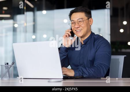 Successful Asian man smiling and talking on the phone, businessman in a shirt working inside the office using a laptop. Stock Photo