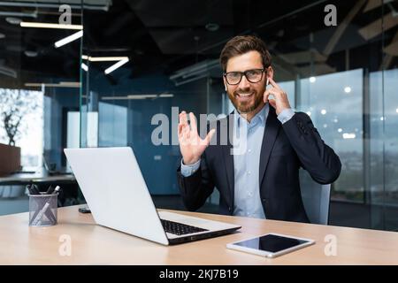 Portrait of a successful businessman inside a modern office, a man in a business suit uses earphones for a video call and remote communication, an investor looks at the camera and uses a laptop. Stock Photo