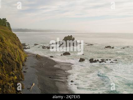 Sea coast. White foamy waves beat against the shore, large stones, boulders in the water. high hill on the shore, covered with moss. A mountain range Stock Photo