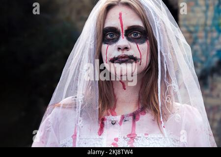 Portrait of zombie woman in wedding dress with veil and stage makeup looking into camera. Girl applied white makeup and blood stains on her face Stock Photo
