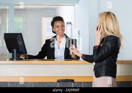 hotel receptionist passing keys to female client talking on smartphone Stock Photo
