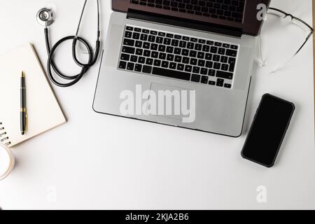 Composition of stethoscope with laptop, smartphone, pen and notebook, on white with copy space Stock Photo