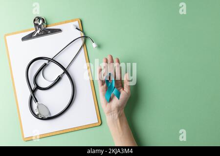 Clipboard, stethoscope and hands holding blue ovarian cancer ribbon on green background, copy space Stock Photo