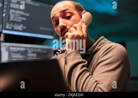 App developer talking on landline phone call after hours, answering office cord telephone to program code on terminal window. Database admin using phone line to develop new interface. Stock Photo