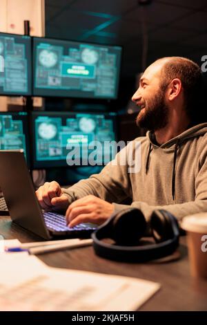 Male hacker stealing computer data from server, wearing hoodie and doing cyberterrorism with multiple screens. Server hacking and cyberattacks using virus to infect network system. Stock Photo