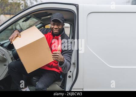 Young adult black delivery man getting out of white van with carboard box parcel looking into camera smiling and showing thumbs up. Horizontal shot. High quality photo Stock Photo