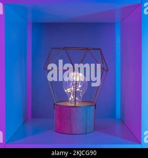 A lamp with a metallic gold geometric lampshade on a wooden stand with an LED lamp in neon light in a white rack. Stock Photo