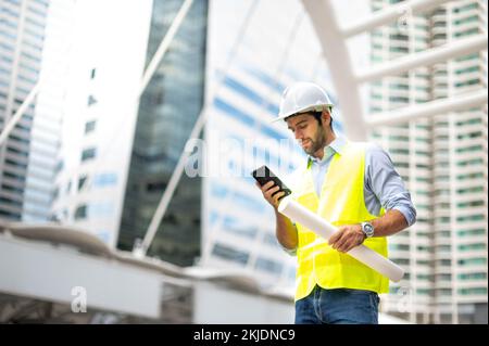 Caucasian man engineers use a smartphone for talking, wearing a yellow vest and a big hard hat, and the other hand holding the white floor plan in the site Stock Photo