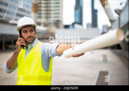 Caucasian man engineers use a smartphone for talking, wearing a yellow vest and a big hard hat, and the other hand holding the white floor plan in the site Stock Photo