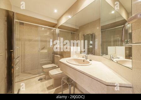 Bathroom with marble countertops, many mirrors, walk-in shower with hinged glass doors, and light brown toilets Stock Photo