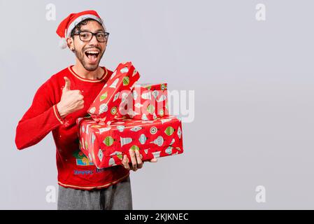 Smiling guy in christmas hat holding christmas gift boxes giving thumbs up. Friendly man in christmas clothes holding gift boxes, Christmas man holdin Stock Photo