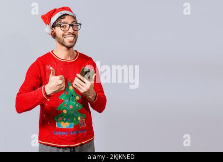 Smiling guy in christmas hat using cellphone with thumb up. Smiling young man in christmas hat using cellphone and giving thumbs up. People in santa h Stock Photo