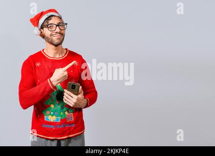 Christmas man holding phone pointing to the side. Smiling young man in christmas hat with cellphone pointing at promo. Christmas man concept with cell Stock Photo