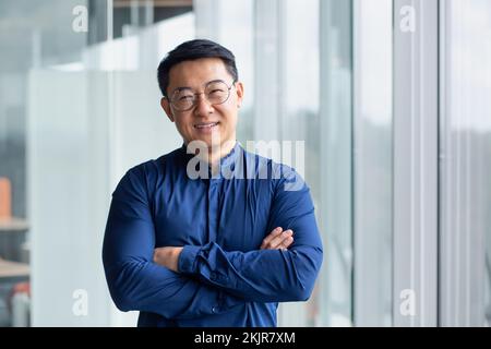 Portrait of successful mature boss, senior businessman in glasses Asian looking at camera and smiling, man with crossed arms working inside modern office building. Stock Photo