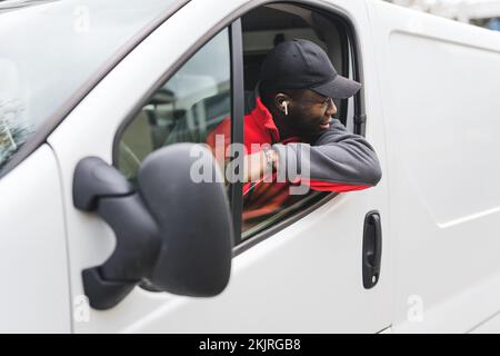 Young adult black delivery guy wearing work unifrom driving white van leaning out the window to look behind. Horizontal shot. High quality photo Stock Photo
