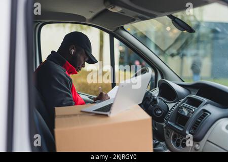 Black young adult delivery guy sitting inside white van signing papers and working on laptop computer. Horizontal shot. High quality photo Stock Photo