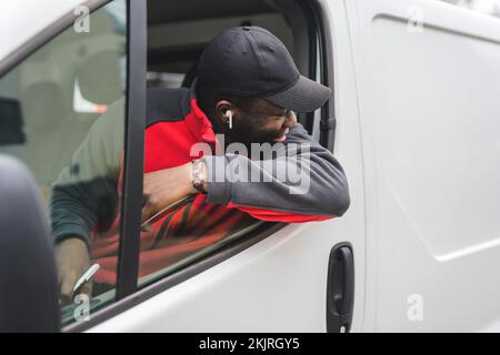 Black young adult delivery man sitting in driver seat of white van leaning out the window smiling looking away from camera. Horizontal shot. High quality photo Stock Photo