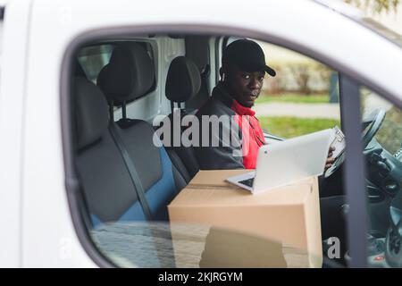 Young adult black delivery guy sitting inside white van filling out papers and working on laptop looking at camera through the window. Horizontal shot. High quality photo Stock Photo