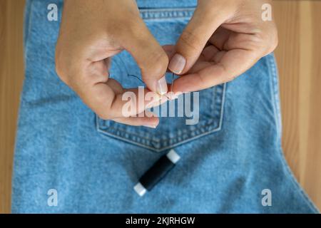 Hands of a seamstress threading a needle on the background of jeans Stock Photo