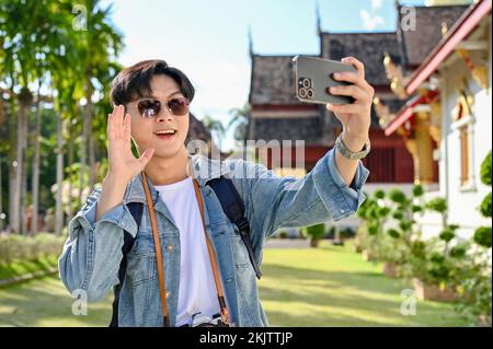 Handsome and cheerful young Asian male traveler wearing sunglasses, taking selfie or making a video vlog with his smartphone in the beautiful Thai's t Stock Photo