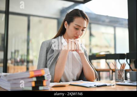 Professional and thoughtful millennial asian businesswoman concentrating on her tasks, analyzing financial data on report, working at her desk. Stock Photo