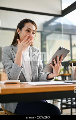 Portrait, Shocked and excited young Asian businesswoman looking at her tablet screen, receiving an unexpected good news. Stock Photo