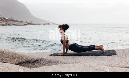 Woman exercising on a mat by ocean. Slim female doing stretching pose outdoors at sunset. Stock Photo