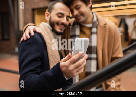 smiling gay man embracing bearded boyfriend using mobile phone outdoors on blurred background,stock image Stock Photo