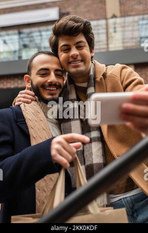 smiling gay man embracing bearded boyfriend with shopping bags while taking selfie on blurred mobile phone,stock image Stock Photo