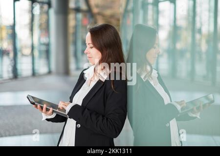 Smiling businesswoman using touchscreen of tablet computer in city leaning on glass wall Stock Photo
