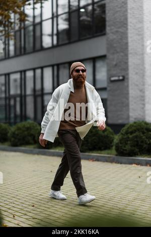 full length of smiling hipster in beanie hat and trendy sunglasses walking in shirt jacket near modern building,stock image