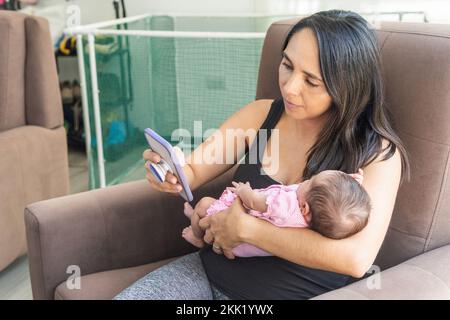 Mature woman with her newborn baby in her arms working at home from her cell phone Stock Photo