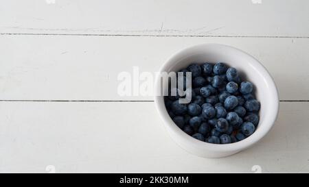 close view of a small white ceramic bowl filled with fresh blueberries. Placed on top of a white wooden table Stock Photo