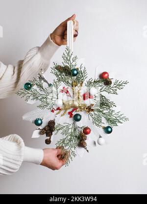 Close-up woman holding snowflake shape Christmas wreath with reindeer White cedar base and cones, no waste festive decoration in female hands. Green Stock Photo