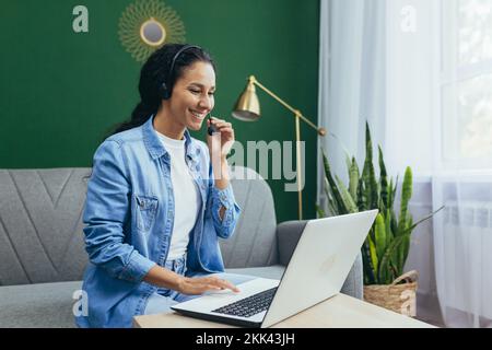 A young, beautiful, smiling Latin American woman blogger conducts a webinar, records a podcast, conducts an online broadcast. Sitting at home on the sofa in headphones with a laptop. Stock Photo