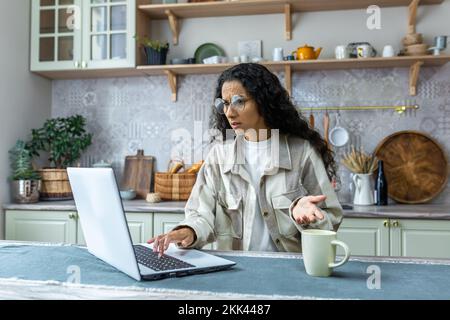 Young hispanic female freelancer in glasses can't work on laptop at home. The lights are off, the deadlines are on. She sits confused and angry in the kitchen at the table, spreading her hands. Stock Photo