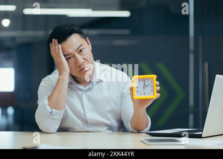 Exhaustion at work, burnout. Tired young man Asian businessman, sitting in the office, holding a watch in his hands. falls asleep, rests his head on his hand. Waiting for the end of the working day. Stock Photo