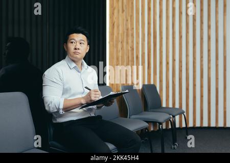 A serious young Asian man fills out a questionnaire for the reception desk, sits in the lobby of an office, hotel, hospital. He holds a folder with papers in his hands, looks to the side. Stock Photo