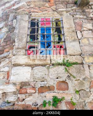 Abandoned weathered old stone bricks wall with corroded antique grunge wrought iron small window with multi vibrant colored squares, in an old village Stock Photo