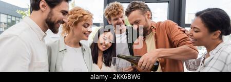 cheerful businessman pouring champagne near smiling interracial colleagues having corporate party in office, banner Stock Photo