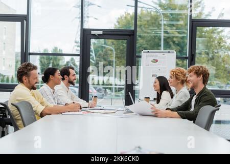 multiethnic corporate team sitting at conference table near flip chart with infographics Stock Photo