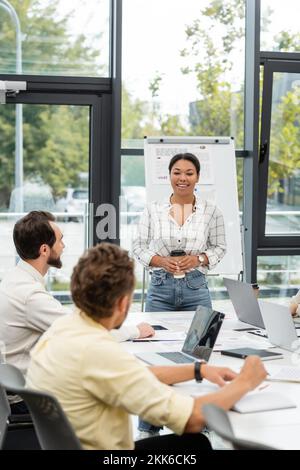 smiling multiracial woman with coffee to go talking to colleagues sitting at conference table Stock Photo