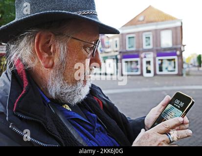 Elderly man with hat is checking his smartphone. He is in the middle of the historical town of Coevorden in the Netherlands Stock Photo