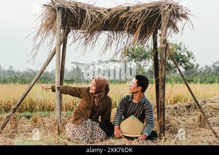 A couple of asian boy and girl sitting in a hut in traditional Javanese clothes in a rice field with plants and blue sky background Stock Photo