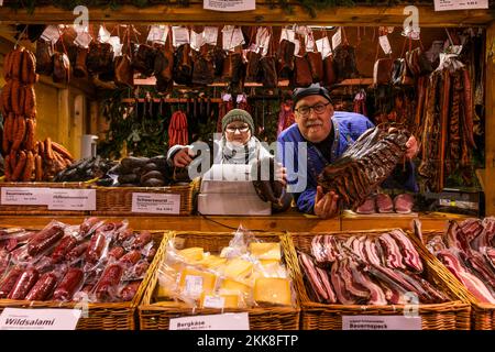 Breitnau, Germany. 25th Nov, 2022. Sandra Kästner and Stefan Gutscher stand in their booth holding black sausage and Black Forest bacon in their hands at the Christmas market in Ravennaschlucht. After the Christmas market in the Ravennaschlucht had to be cancelled due to the Corona pandemic in 2020 and was only able to open for a few days in the following winter of 2021, this year visitors are allowed to visit the Christmas market under the viaduct of the Höllentalbahn on all four Advent weekends. Credit: Philipp von Ditfurth/dpa/Alamy Live News Stock Photo