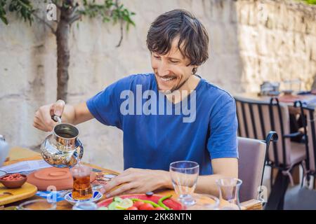 Man eating turkish breakfast. Turkish breakfast table. Pastries, vegetables, greens, olives, cheeses, fried eggs, spices, jams, honey, tea in copper Stock Photo