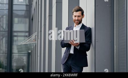 Successful man standing in city near office building typing on laptop professional businessman architect designer develops online project browsing Stock Photo