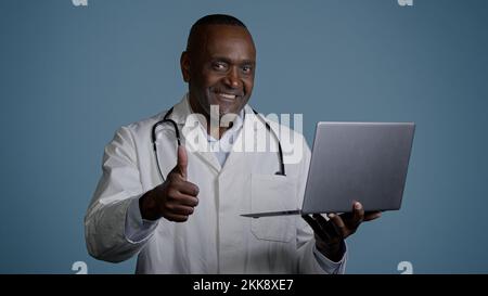 Mature man stands in studio on gray background professional doctor in medical gown checks tests using laptop consults patient online showing thumb up Stock Photo