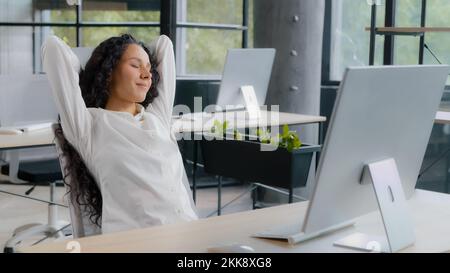 Relaxed woman manager taking break at workplace holding hands behind head resting after completing work sitting at desk dreaming with closed eyes Stock Photo